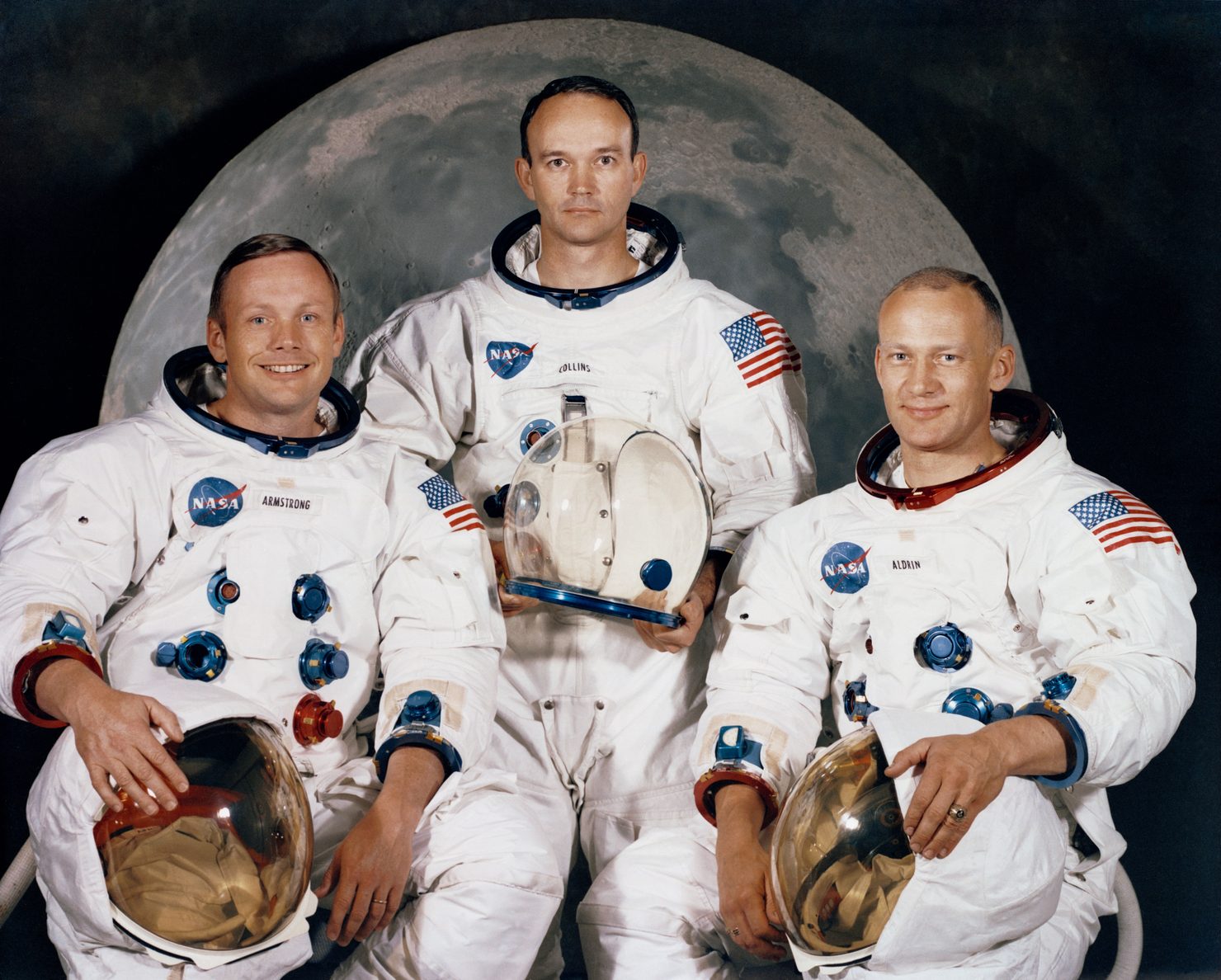 The Apollo 11 crew in white pressure suits: Neil Armstrong, Michael Collins holding his helmet, and Buzz Aldrin.