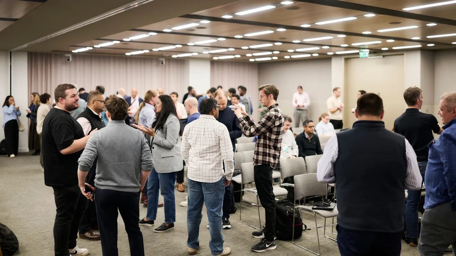 Developers talking in small groups at one of the bank’s two-day coding-agent workshops, with rows of chairs in a conference room.